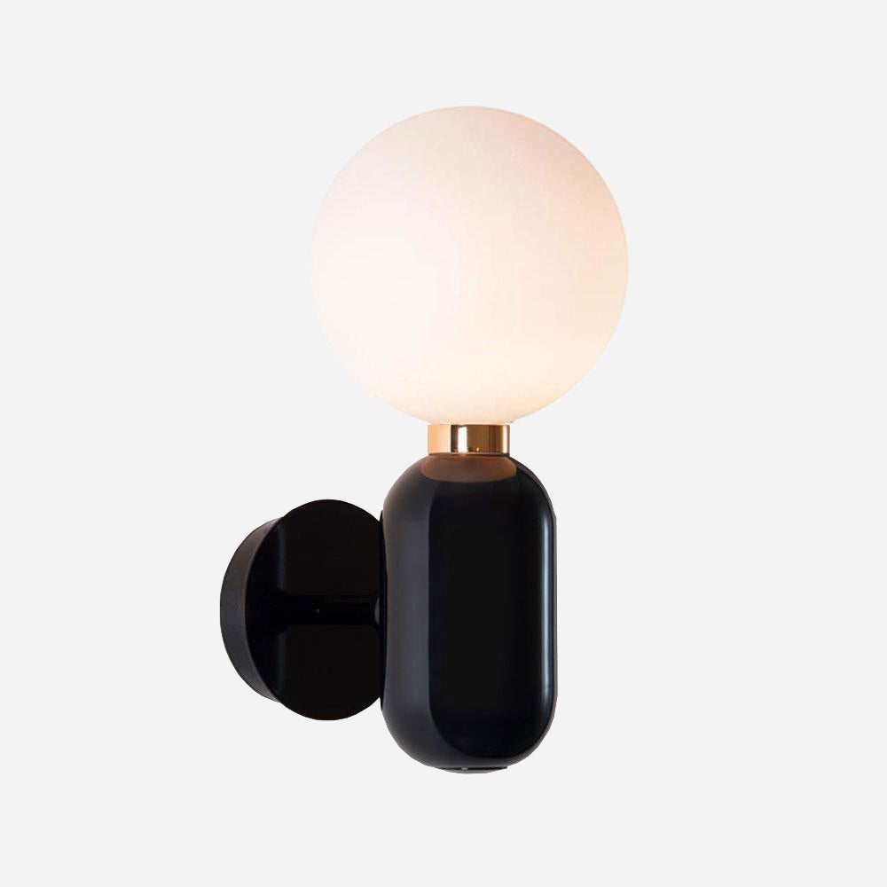 The Pallo Wall Light - Black showcases a sophisticated black finish complemented by an opal glass diffuser, featuring a round frosted globe gracefully perched on a stylish cylindrical base with a gold accent. When mounted on a light-colored wall, it emits a soft, warm glow that seamlessly blends modern elegance with cozy ambiance.