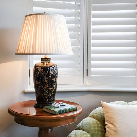 A Black Birds Table Lamp - Black & Gold with a floral design sits on a round wooden side table. Next to it is a stack of books with a pair of glasses on top. The scene, reminiscent of traditional Chinese craftsmanship, is framed by white window shutters and a partially visible patterned green armchair.