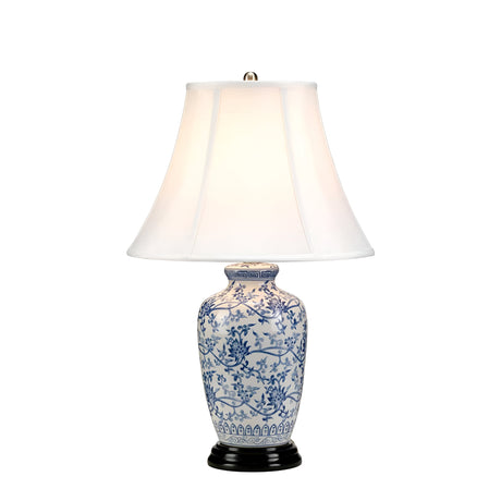 The Blue Ginger Jar Table Lamp - Blue & White features a decorative design with a white fabric shade and a classic ceramic base adorned with blue floral patterns. It rests on a small black wooden stand, and when lit, it casts a warm glow that highlights its elegant Chinese craftsmanship.