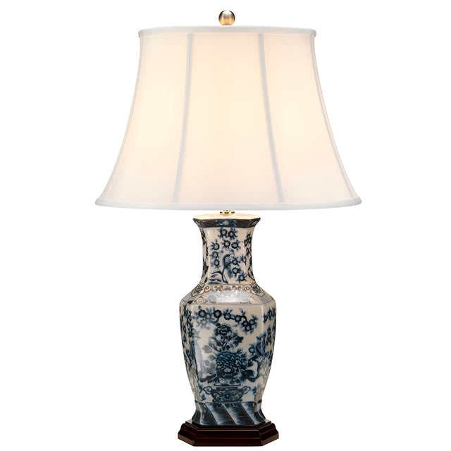 The Blue Hex Table Lamp - Blue & White showcases a porcelain base adorned with blue and white floral patterns, echoing traditional Chinese craftsmanship. It is fitted with a white bell-shaped fabric shade, sits on a wooden base, and is finished with a small finial on top.