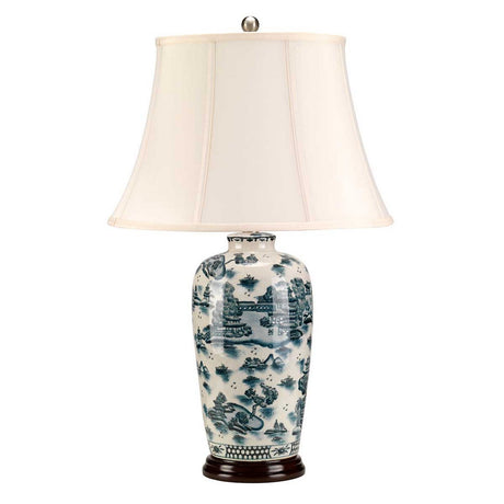 The Blue Traditional Table Lamp - Blue & White features a ceramic base with detailed landscape designs, evoking the classic look of Chinese porcelain. It is completed with a white flared lampshade, a small metallic finial, and a dark round stand that adds to its cultural elegance.