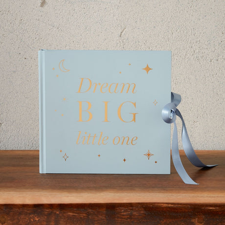 Pale blue baby photo album with gold "Dream BIG little one" text, moon and star details, and a grey ribbon tie, standing upright on a wooden shelf.