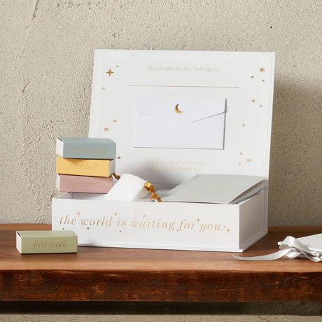 White "Our Little One" keepsake box with gold text and stars, open to show a notebook, pouch and envelope, with pastel first tooth, curl and words boxes on a wooden shelf.