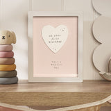 1st Birthday Heart Framed Wooden Plaque in white frame with pink panel, raised white heart reading "on your first birthday, have a magical day", shown on a nursery shelf.