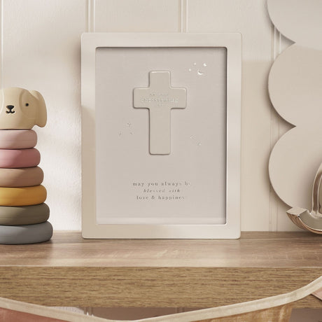 Christening Cross Wooden Framed Plaque, 17cm, white frame with beige panel, raised cross, stars and moon detail, shelf display beside nursery toys.