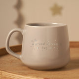 Grey stoneware mug with silver "a grand adventure begins" lettering, stars and crescent moon design, rounded handle, shown on a wooden surface.
