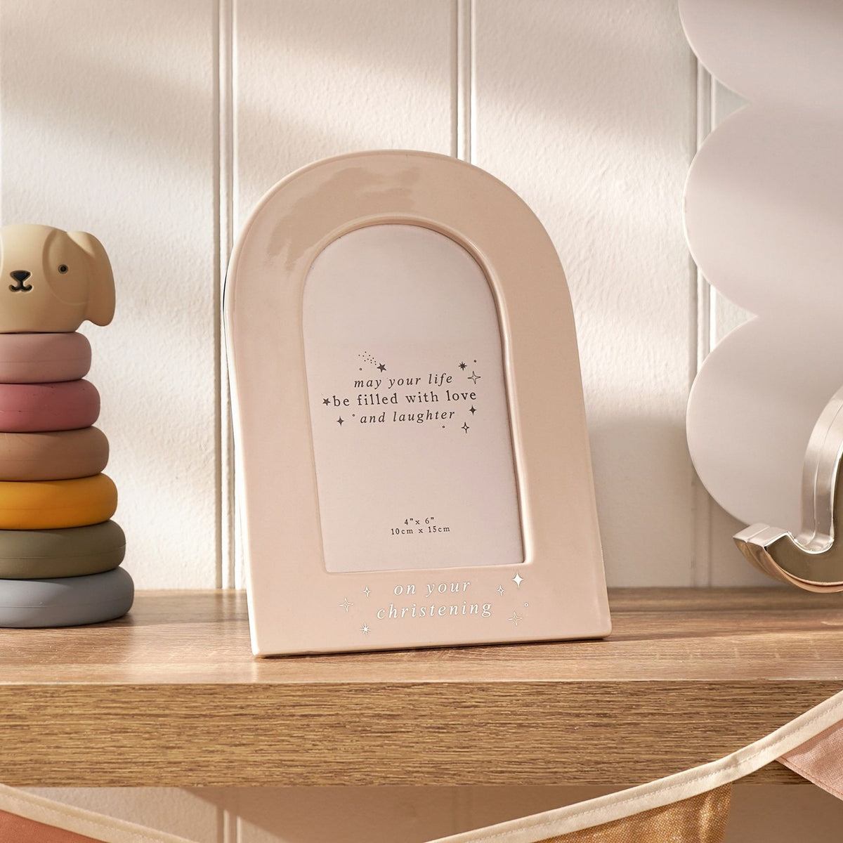 Cream ceramic arch photo frame for a 6 x 4in print, with "on your christening" in silver foil and star details, shown on a wooden shelf beside toys.