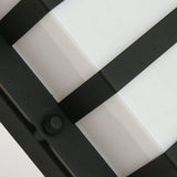 A close-up view of the Boden Outdoor 1 Light Wall Light showcases its black metal grid structure with three intersecting bars against a smooth, white, opaque surface. The design elegantly highlights the Art Deco aesthetic, complete with a circular frosted glass element on the left side.