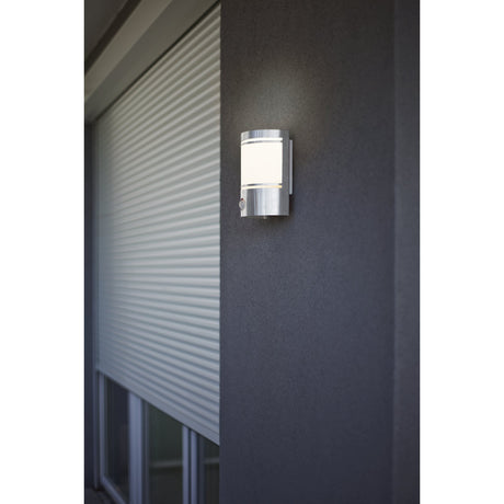 Bonvenne outdoor wall light in stainless steel with motion sensor and white frosted glass shade, mounted on a dark grey exterior wall beside a window.