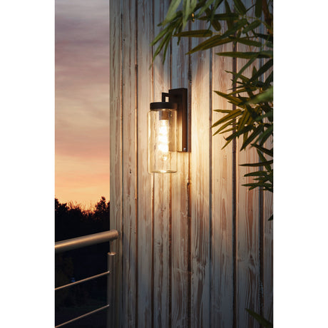 Bovolone black outdoor wall light with clear glass cylinder shade and L-shaped arm, illuminated at dusk on a wooden slatted exterior wall.