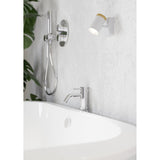 Brelora Wall Spotlight in white steel with a cylindrical head and light wood accent, mounted above a bathroom tub beside a chrome handheld shower and basin tap.