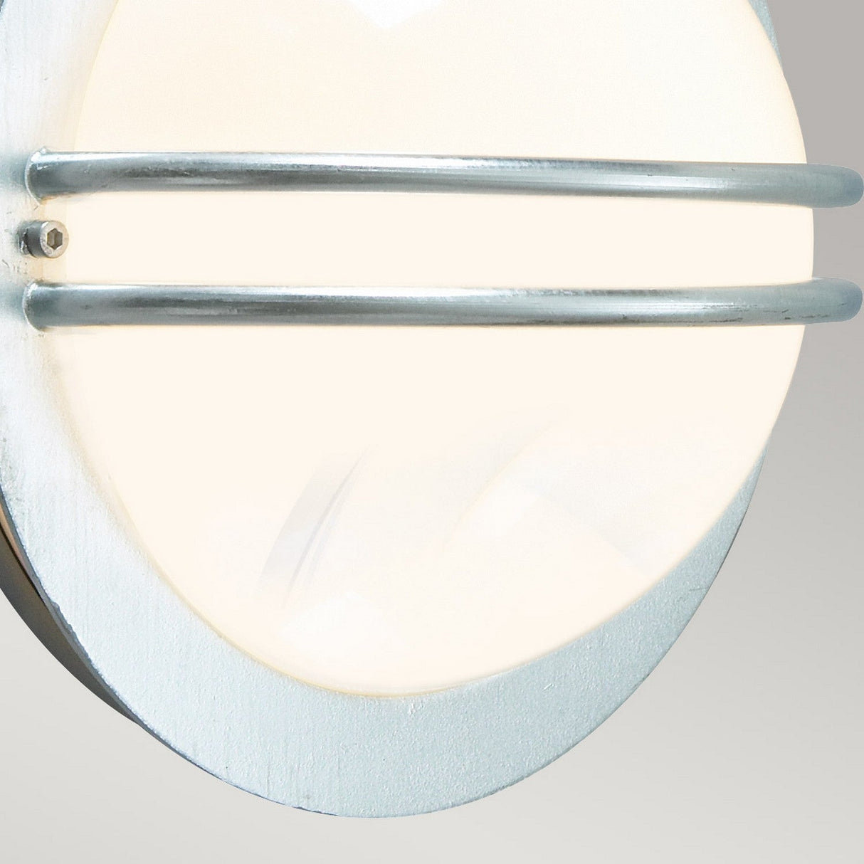 Close-up of the Bremen Outdoor 1 Light Wall Lantern - Silver, a wall-mounted circular fixture featuring a frosted glass cover. It is secured by two parallel metal bars and boasts a modern industrial design with nautical charm. Made from galvanized steel with a metallic finish, it emits a soft white glow.