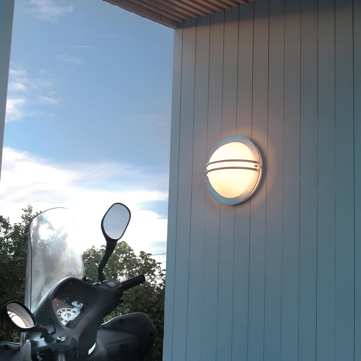 Mounted on a blue exterior wall near the corner of a building is the Bremen Outdoor 1 Light Wall Lantern in silver, exuding nautical charm. Nearby, a scooter equipped with a windshield and mirrors is parked. In the background, green shrubs and a clear sky further enhance the maritime aesthetic.