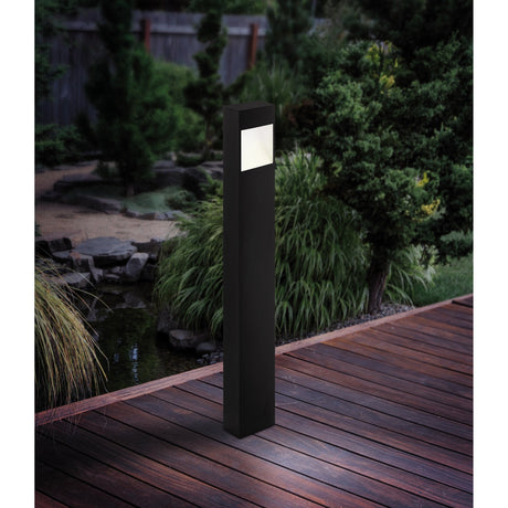 Bresorne LED bollard light with tall anthracite aluminium body and white illuminated window, standing on a wooden deck beside garden greenery at dusk.