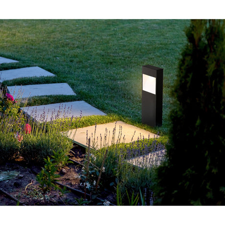Bresorne LED outdoor post light in anthracite with a white-lit window, shown illuminated on a lawn beside a garden path with flowers and paving stones.