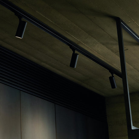 Brillia 1m black surface-mounted lighting track fitted to a ceiling, shown with three cylindrical spotlights attached along its slim rectangular rail.