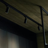Brillia 2m surface-mounted 1-phase lighting track in black, fitted with three cylindrical spotlights along a concrete ceiling beam.