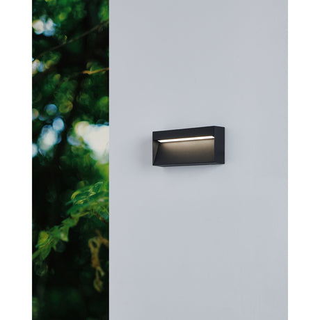 Brolbenne LED Outdoor Wall Light in black aluminium, rectangular design with angled face and integrated LED strip, mounted on a light grey wall near foliage.