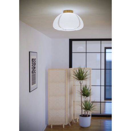 Brosmena flush ceiling light with a ribbed white paper lantern shade and round wooden base, shown illuminated in a room with a plant and woven screen divider.