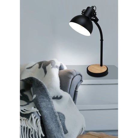 Brosora Table Lamp in black steel with wood-topped round base and curved arm dome shade, shown lit on a bedside table beside a grey armchair with throw.
