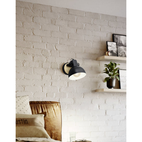 Brosora Wall Light in black and wood, a black steel dome shade on a round wooden backplate, mounted on a white brick wall beside floating shelves in a living room.