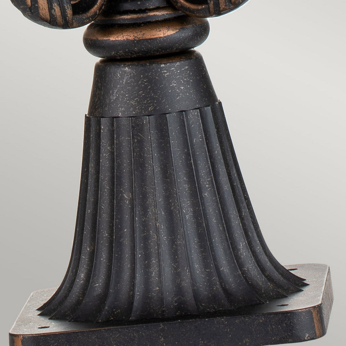 A detailed view of the Baltimore 1 Light Large Pedestal Lantern showcases a weathered bronze base with a textured, fluted design. The surface features a subtle metallic sheen with speckled aged accents for an authentic vintage appeal. Echoing classic outdoor lanterns, the base gracefully tapers upward and rests on a square platform.