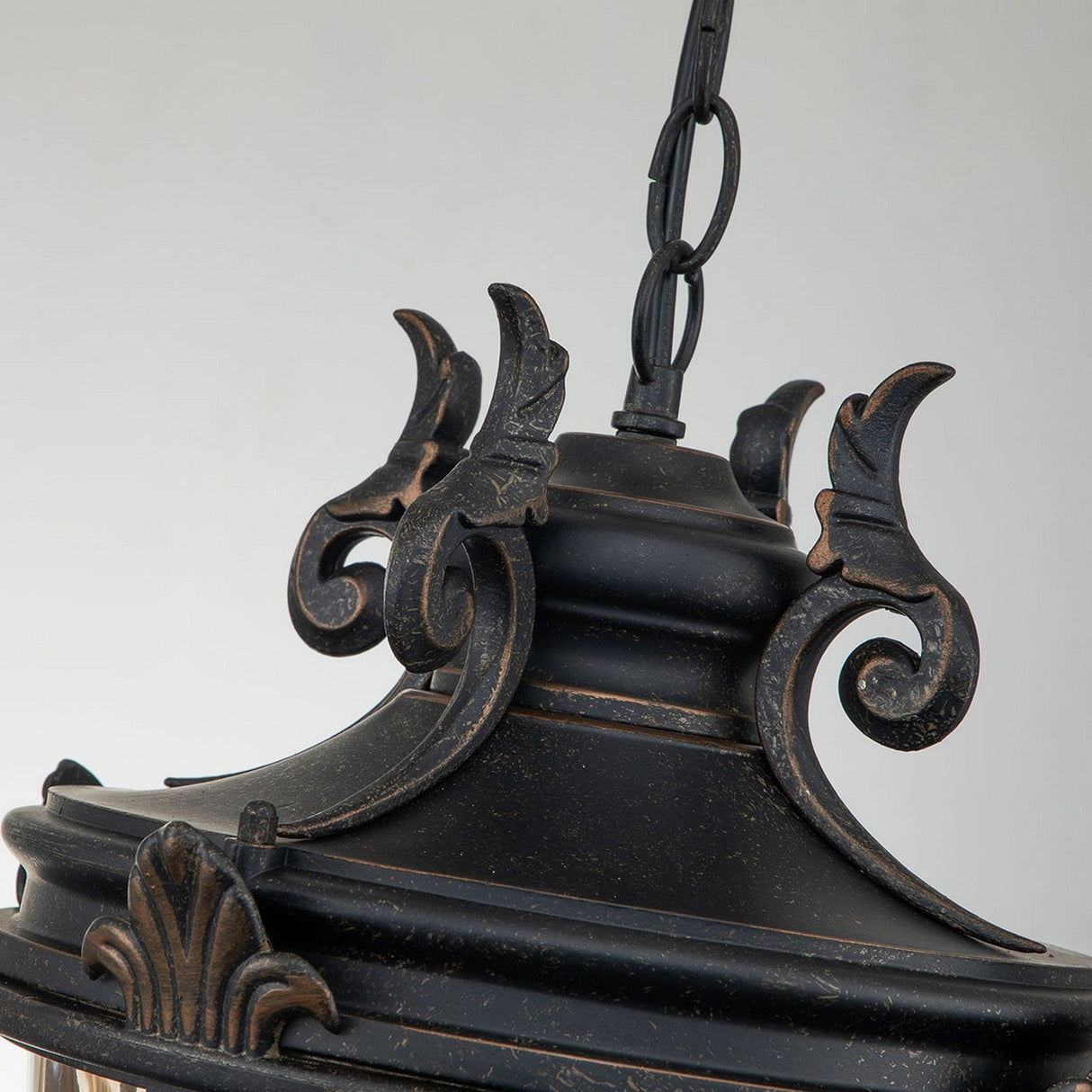 Close-up of an ornate black hanging lantern showcasing intricate metalwork and a weathered bronze finish. Reminiscent of the Baltimore 1 Light Outdoor Large Chain Pendant, it features curved leaves and a chain for suspension, set against a plain gray background. Ideal for outdoor lighting.