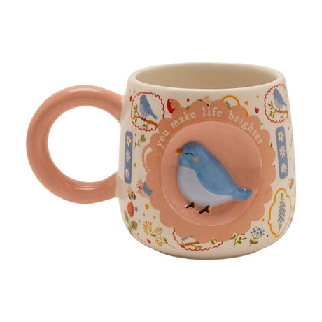Ceramic mug in cream with a pink handle, featuring a raised 3D blue bird on a scalloped medallion reading "you make life brighter," surrounded by a wildflower print, shown on a plain white background.