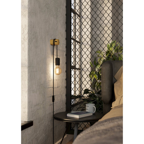 Buddon Wall Light in black and wood, a plug-in single E27 wall light with round wood mount and black cord lampholder, shown lit beside a bed in an industrial-style bedroom.