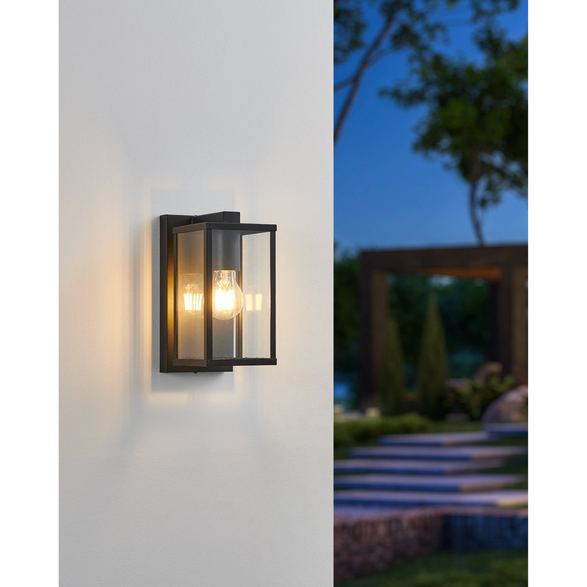 Budrone outdoor wall light in black steel with clear glass panels and visible E27 bulb, mounted on a wall at dusk near a garden setting.