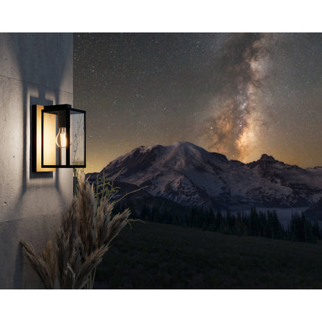 Budrone outdoor wall light with black steel box lantern frame, clear glass panels and brown wood-effect back plate, mounted on a wall at night with mountain scenery beyond.