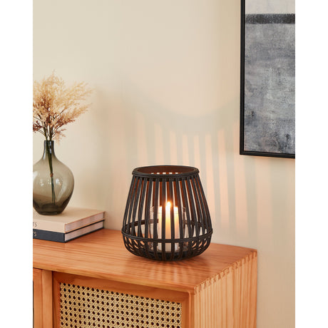 Bunawan bamboo candle lantern in black, a rounded slatted design with a clear glass insert holding a lit candle, displayed on a wooden sideboard beside a vase and books.