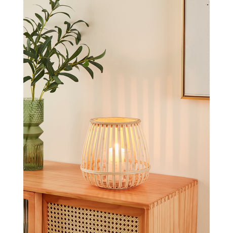 Bunawan bamboo candle lantern with natural slatted sides and glass holder, lit candle glowing inside, shown on a wooden sideboard beside a green vase and plant.