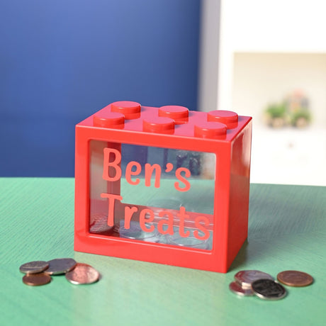 Red plastic building-block money box with clear window reading "Ben's Treats" in red letter stickers, shown with loose coins on a green surface.