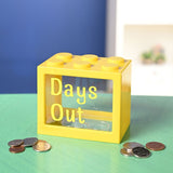 Yellow building-block shaped money box with a clear front window reading "Days Out" in yellow letters, displayed with scattered coins on a green surface.
