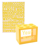 Yellow block-shaped money box with six top studs and a clear window for name stickers, shown with a matching yellow alphabet sticker sheet on white background.