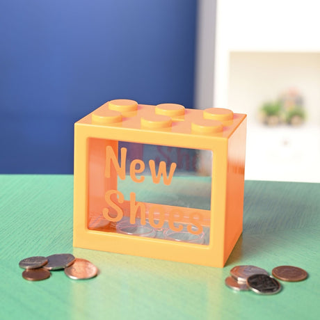 Orange plastic building block-style money box with clear window front, "New Shoes" alphabet stickers applied, shown on a green surface with coins scattered beside it.