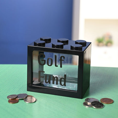 Black plastic building-block style money box with six top studs and a clear front window spelling "Golf Fund" in alphabet stickers, shown with scattered coins on a green surface.