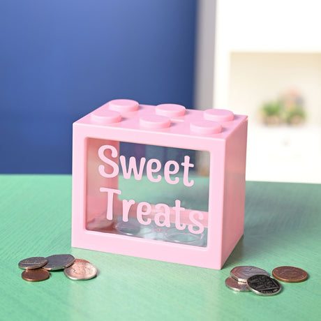 Pink plastic building block-shaped money box with six studs and a clear front window reading "Sweet Treats", shown on a green table with coins.