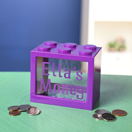 Purple plastic building block-shaped money box with six top studs and a clear window, personalised with alphabet stickers spelling "Ella's Money", shown with coins on a green surface.