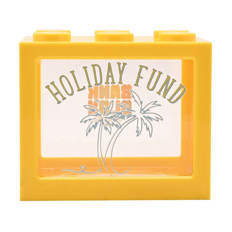 Yellow plastic building block money box with six top studs and a clear front window printed "Holiday Fund" above palm tree outlines, shown on a plain white background.
