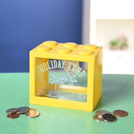 Yellow plastic building block-style money box with six top studs and a clear front window reading "Holiday Fund" above palm tree outlines, shown with scattered coins on a green surface.