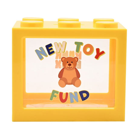 Yellow plastic building block money box with six top studs and a clear front window showing a teddy bear and colourful "New Toy Fund" text, on a plain white background.