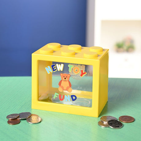 Yellow plastic building block money box with a clear front window showing "New Toy Fund" in rainbow letters around a teddy bear, displayed with coins on a green surface.