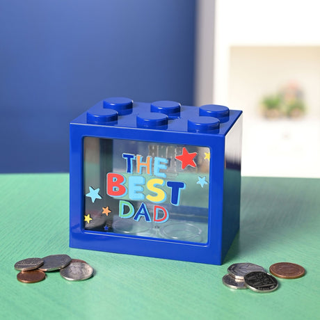 Navy blue plastic building-brick money box with six top studs and a clear window reading THE BEST DAD in colourful lettering with stars, shown on a green surface beside loose coins.