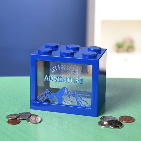 Navy blue building block-shaped money box with six studs, coin slot, and clear front window reading Let's Go on an Adventure above a mountain design, on a green surface with scattered coins.