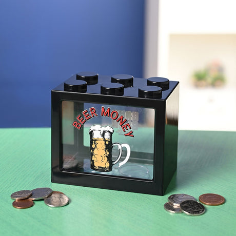 Black plastic brick-shaped money box with six top studs and a clear front window printed "BEER MONEY" above a beer mug, shown on a green surface with scattered coins.