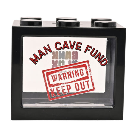 Black plastic building block-shaped money box with six round studs on top and a clear front window reading "MAN CAVE FUND" and "WARNING KEEP OUT" in red text, shown against a plain white background.
