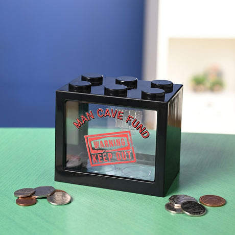 Black plastic Man Cave Fund money box shaped like a building block with six top studs and a clear window reading "Warning Keep Out", shown with loose coins on a green surface.