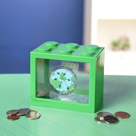 Green plastic building block money box with clear front panel printed with a globe, aeroplanes, and "Saving for an adventure" text, shown with loose coins on a green surface.